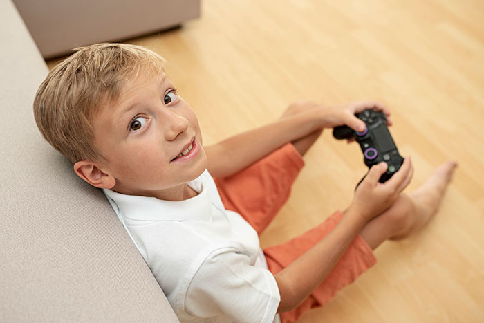 Boy holding a game controller sitting on the floor, looking up with a smile, in a bright room with wooden flooring.