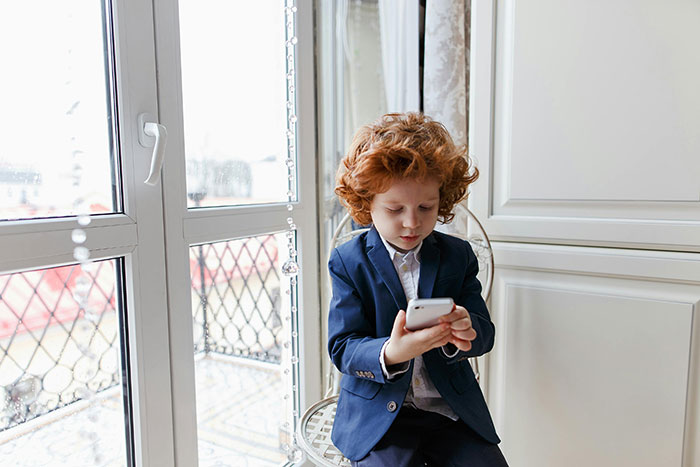 Red-haired boy in a navy blazer sitting by a window, focused on his smartphone in a bright indoor setting.