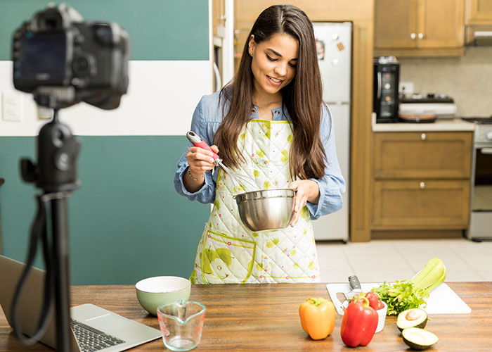 Woman in apron preparing food in kitchen for tradwife lifestyle video recording with camera and laptop nearby