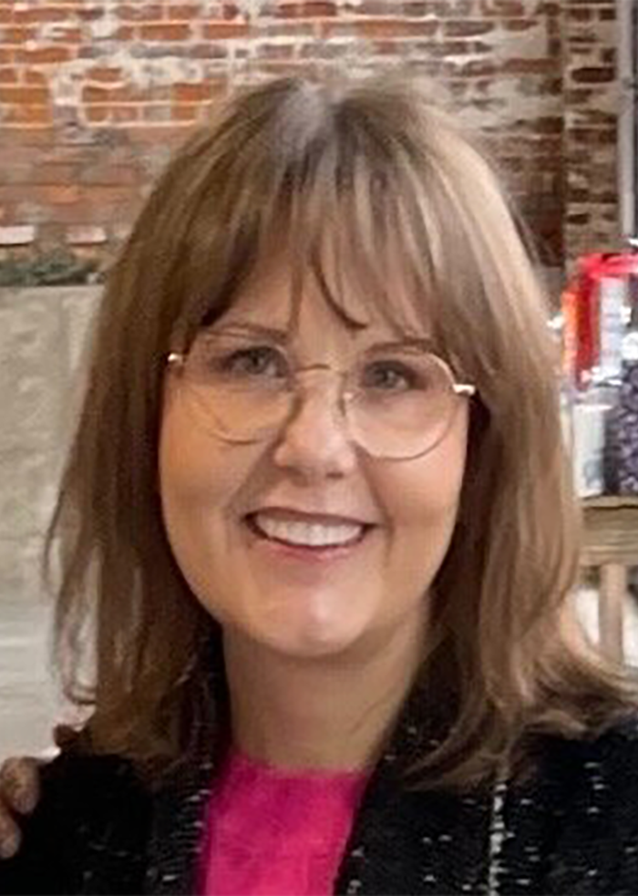 Woman smiling after facelift, wearing glasses with shoulder-length brown hair and a bright pink top in casual setting.