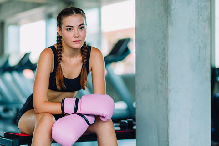 Young woman with pink boxing gloves sitting in gym, focused and determined, related to gym stalker incident story.