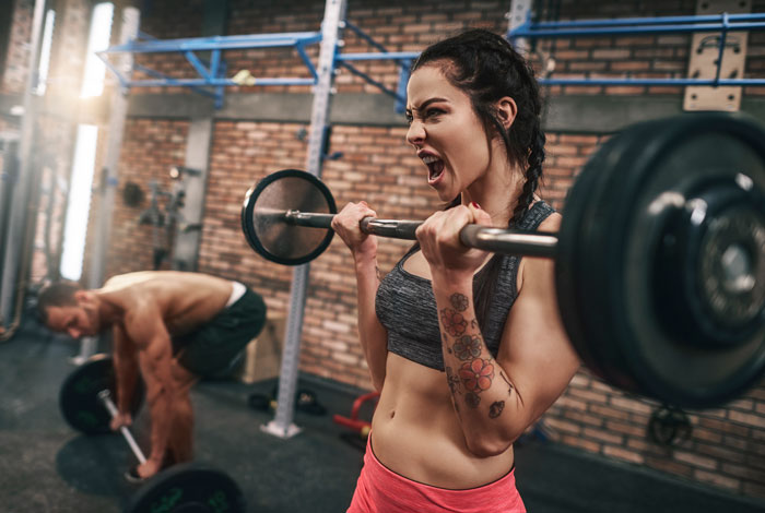 Woman lifting weights in a gym with intense focus while a man avoids unwanted attention nearby.