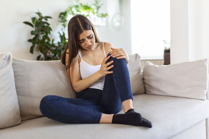 Young woman sitting on a couch holding her knee in discomfort, representing postpartum OCD emotional and physical struggles.