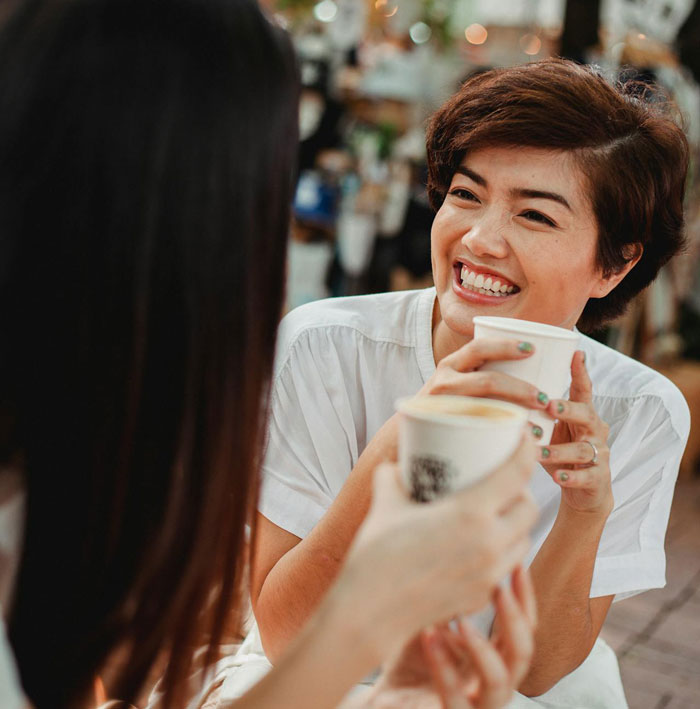 Two women talking and holding coffee cups, illustrating a woman listed as emergency contact refusing to pick up kid.