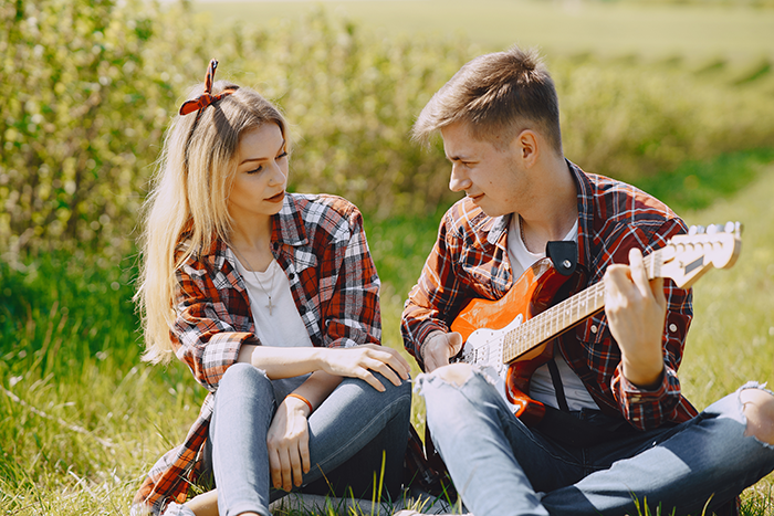 Young woman and man sitting on grass outdoors, with the woman listening as the man plays an electric guitar, illustrating woman move Alaska alone.