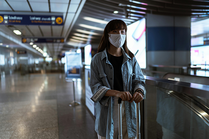 Woman move Alaska alone, standing at an airport with a suitcase, wearing a face mask and casual denim jacket, ready to travel.
