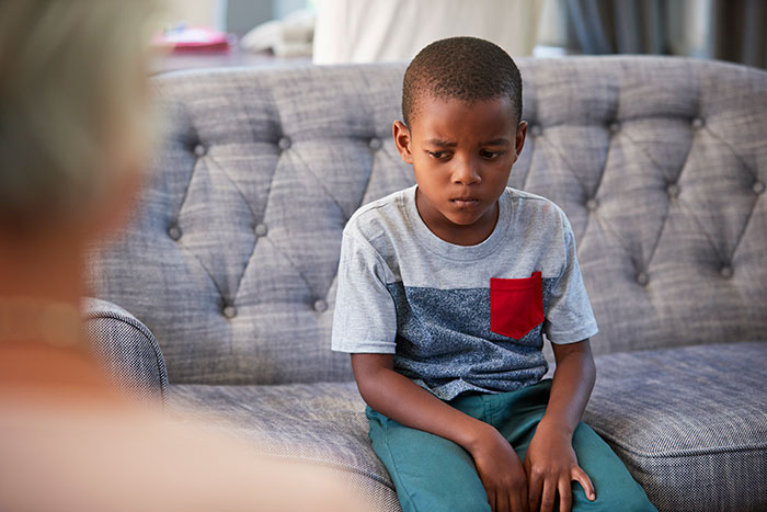 Young boy sitting on a couch looking upset while a babysitter refuses to watch him, raising discrimination concerns.