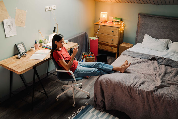 Young woman in casual clothes relaxing in bedroom, focused on her phone, with cozy lighting and a study desk nearby.
