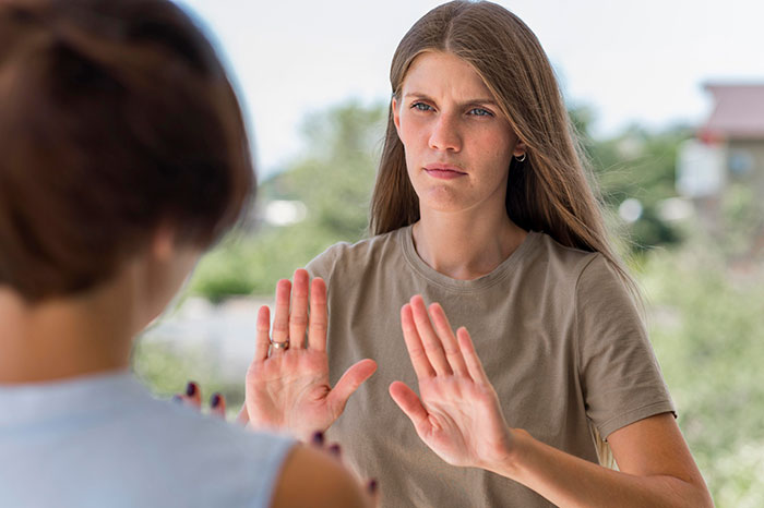 Young woman with serious expression holding hands up in a stop gesture during a prank involving girlfriend&rsquo;s sister outdoors.