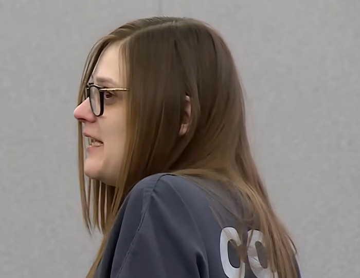 Woman in courtroom wearing glasses and prison uniform, involved in case of killing boyfriend over tampon discovery.