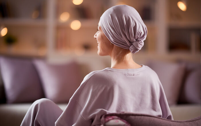 Woman in a light purple headscarf and sweatshirt sitting indoors, reflecting on health and tobacco avoidance advice from oncologists.