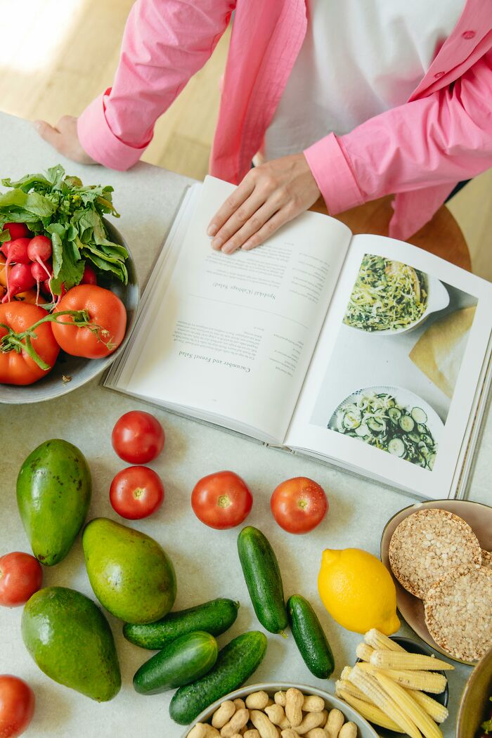 Person in pink shirt reading a cookbook surrounded by fresh vegetables, illustrating disrespectful partner’s family stories topic.