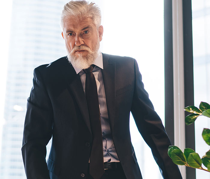 Older man with white beard in a black suit standing by a window, symbolizing employee working 30 hours overtime at office.