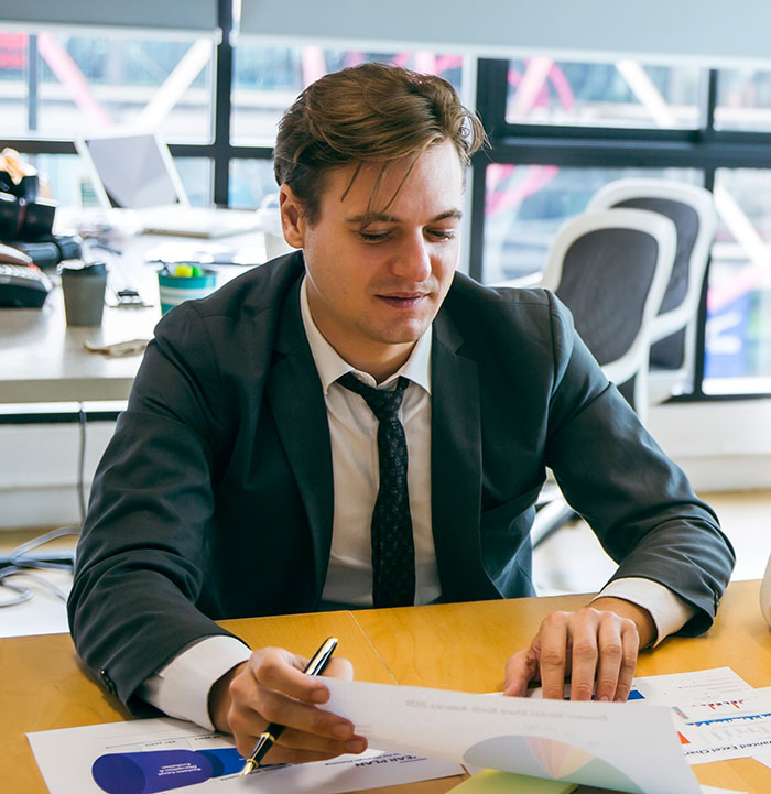 Employee working overtime reviewing documents at office desk, focusing on reports with charts and data analysis.