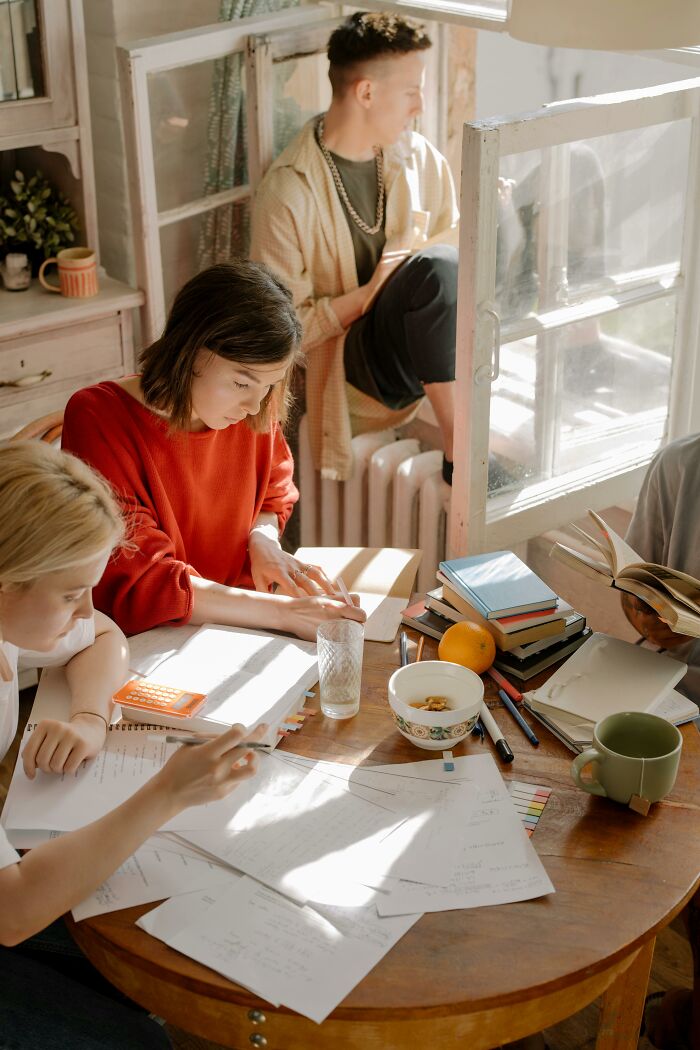 Group of young people studying together at a table with books and papers, showing awkward moments of kindness.