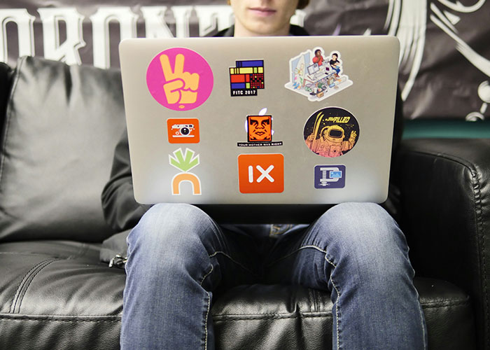 Person sitting on a black leather sofa using a laptop with colorful stickers, illustrating workplace behavior issues.