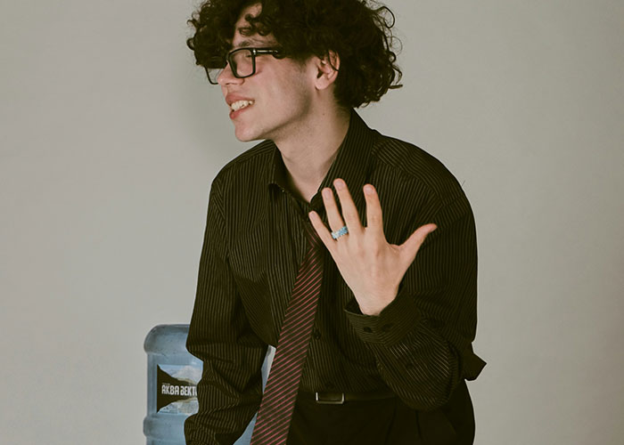 Young man with curly hair and glasses wearing a striped shirt and tie, showing hand gesture in a casual office setting.