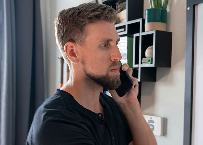 Man with beard and short hair talking on phone in a home office, showing poor work behavior and frustration.