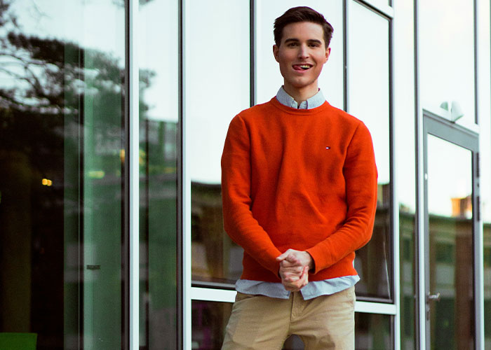 Young man outside office building, wearing orange sweater and beige pants, showcasing unusual work behavior and attitude.