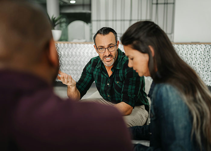 Man in green plaid shirt discussing workplace behavior with two colleagues in a modern office setting.