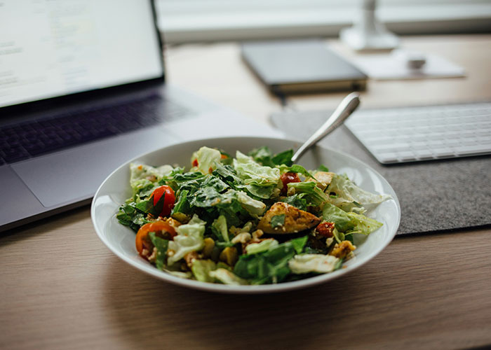 Fresh salad in a white bowl on a desk near a laptop and keyboard showing a typical work environment lunch setting.
