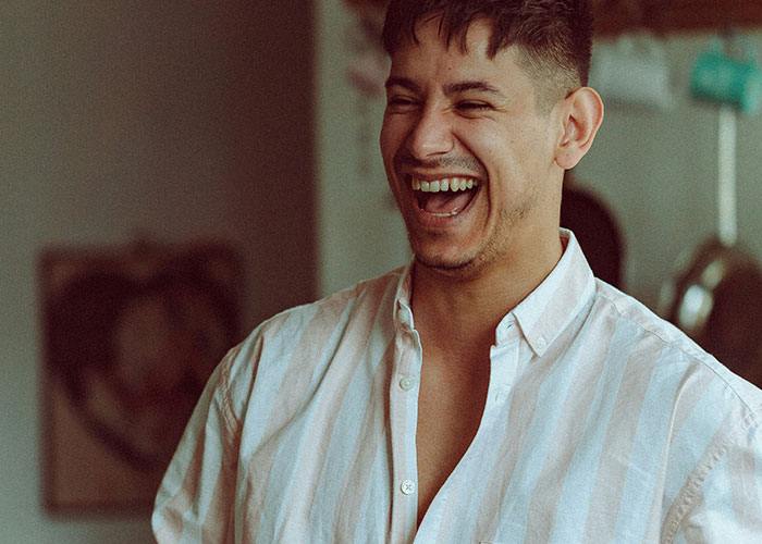 Young man in a casual striped shirt laughing heartily indoors, illustrating people who don’t know how to behave at work.
