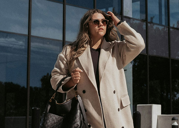 Woman in a beige coat and sunglasses standing outside a modern office building, reflecting workplace behavior issues.