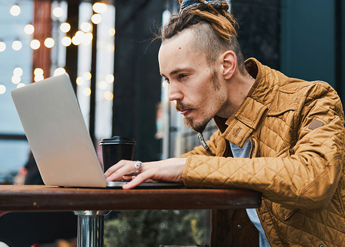Man with dreadlocks focused on laptop screen at outdoor cafe, illustrating people who don’t know how to behave at work.