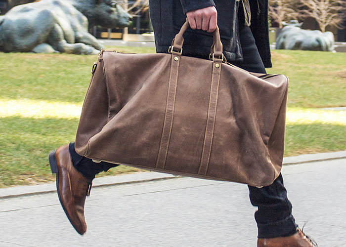 Person walking on sidewalk carrying large brown leather bag, illustrating people who don’t know how to behave at work.