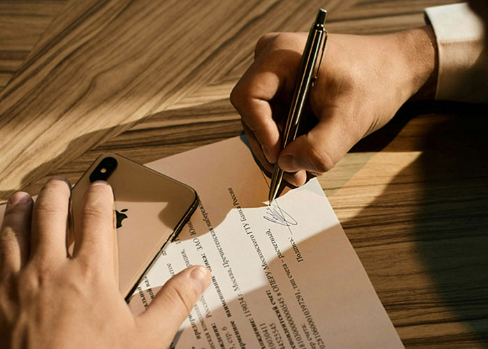 Person signing a document with a pen on a wooden table, next to a smartphone, illustrating workplace behavior.