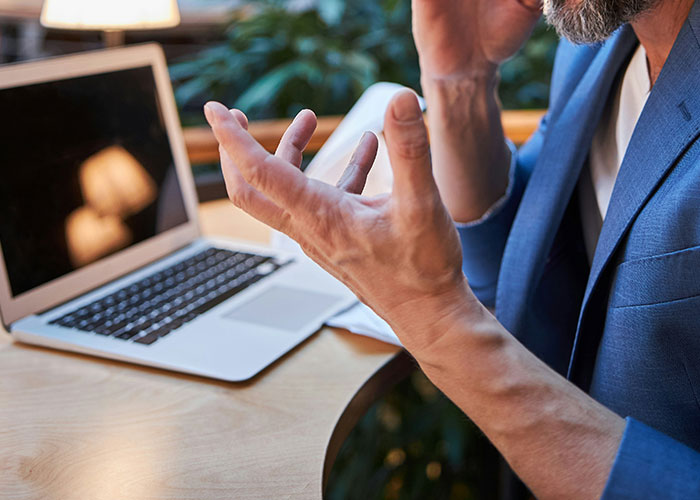 Man in blue blazer gesturing in frustration while sitting at desk with laptop, illustrating poor behavior at work.
