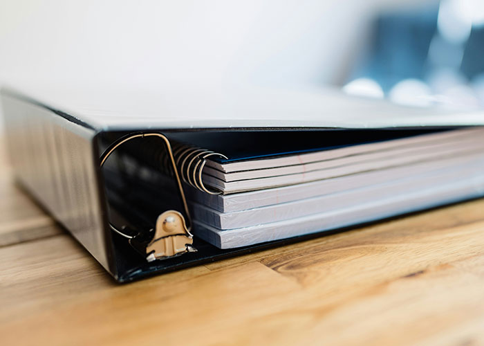 Close-up of a black office binder filled with paper on a wooden desk, symbolizing workplace behavior and organization.