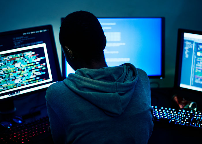 Person working late at a computer with multiple screens displaying code in a dark office environment, illustrating work behavior.