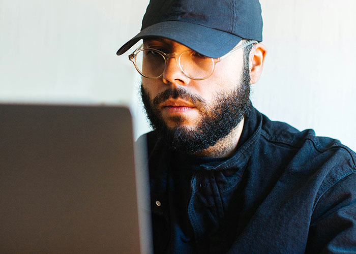 Man wearing a cap and glasses working on a laptop, illustrating people who don’t know how to behave at work.