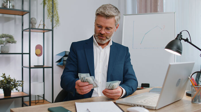 Man in business suit counting cash at desk with laptop, illustrating finance bros profession stereotype. - 8