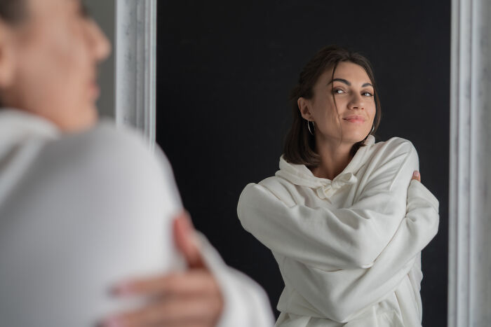 Young woman hugging herself and smiling in front of a mirror, representing dark truths of human behavior and self-perception.
