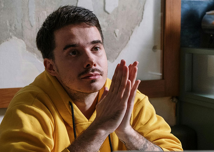 Young man in a yellow hoodie thoughtfully clasping hands, illustrating guy asks ex to move out after breakup concern. - 1