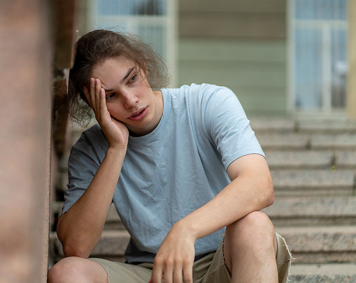 Teen boy sitting on steps looking upset and frustrated about family support and cancer-related head shaving struggles.