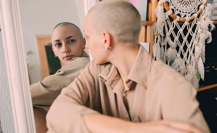 Teen with shaved head looking in mirror, reflecting family upset over not shaving head to support sibling with cancer