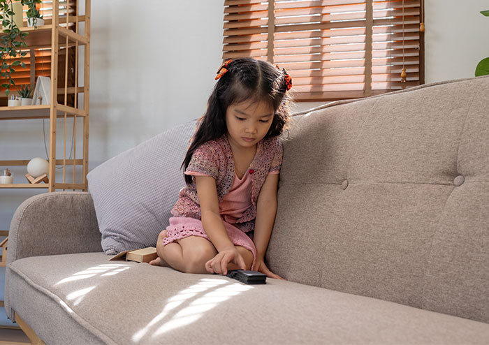 Young girl sitting alone on a couch looking at a remote, depicting a child left alone without babysitting in an emergency. - 1