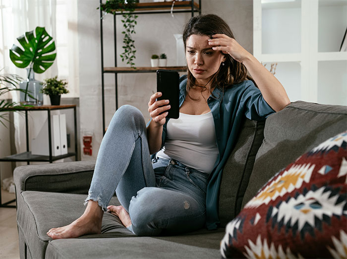 Young woman sitting on a couch looking upset while holding a phone, related to bride refuses to move wedding. - 1