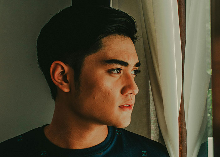 Young man looking out window with concerned expression, possibly upset about a neighbor's farm stand dispute.