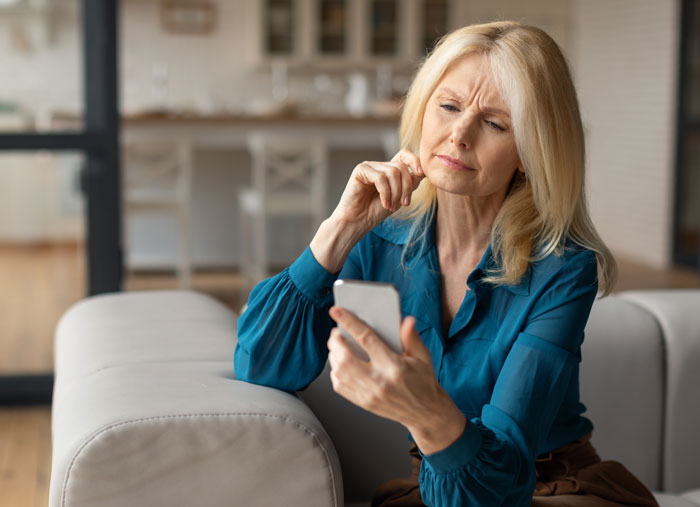 Woman using mil-tracking-app-son on smartphone, sitting on couch in home, looking thoughtful while checking device.