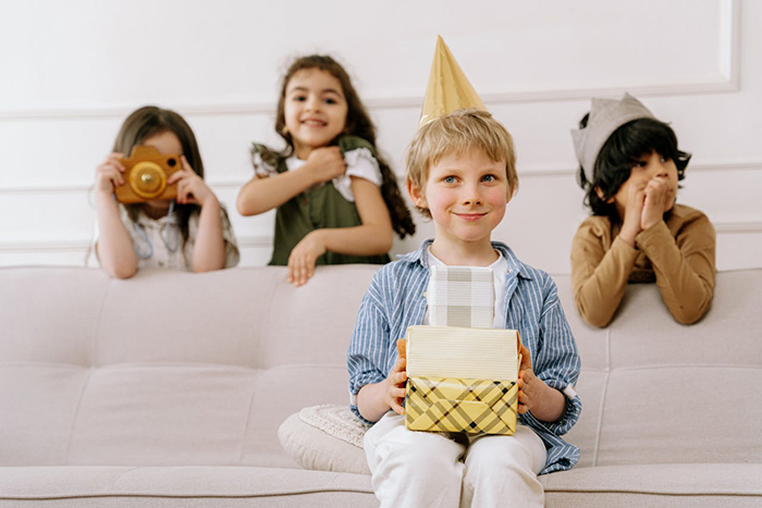 Young boy wearing party hat holding gift boxes while children sit on couch in a playful party scene upset by broken toy incident