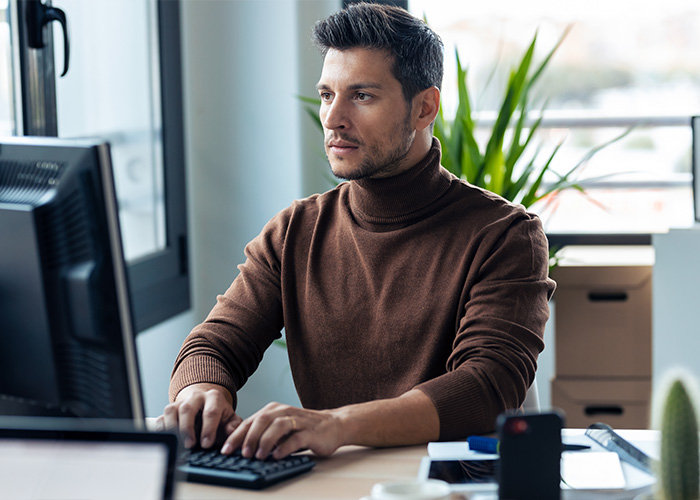 Man in brown sweater typing on keyboard at office desk, relating to employee complaints received by managers and HR.