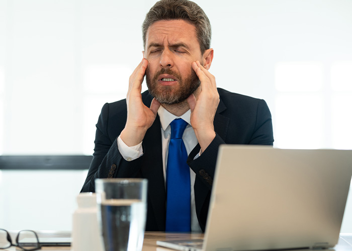 Stressed businessman in suit at office desk rubbing temples, representing ridiculous complaints from employees to HR and managers.