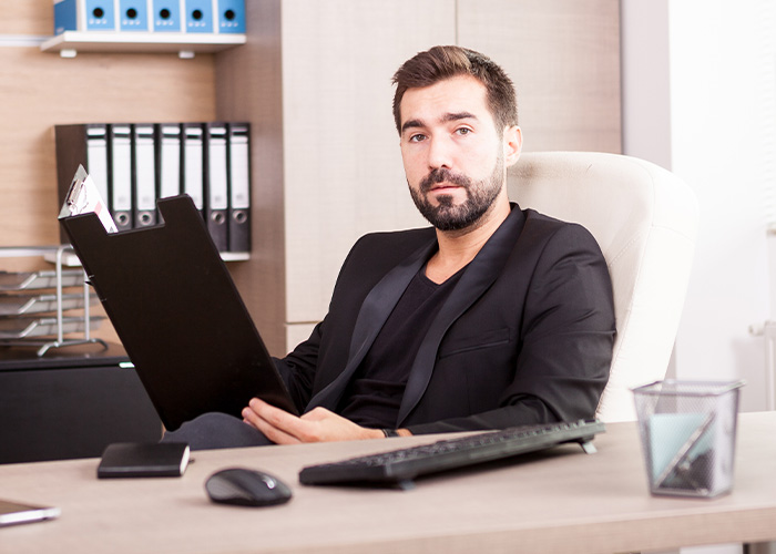Man in black suit sitting at desk with clipboard, representing ridiculous complaints managers and HR receive from employees