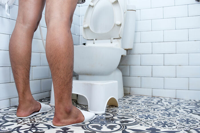 Man standing in bathroom wearing white slippers in front of toilet, illustrating things men find normal but women find bizarre.