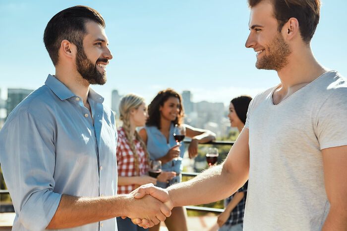 Two men shaking hands outdoors while women socialize in the background, illustrating things normal to men but bizarre to women.