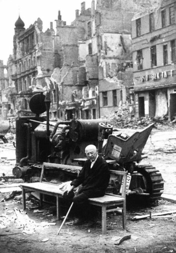 Elderly man with cane sitting on bench in front of machinery amid ruins in a unique old photo from wartime destruction.